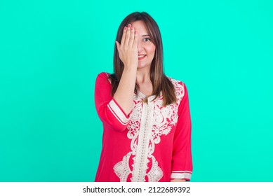 Young Beautiful Moroccan Woman Wearing Traditional Caftan Dress Over Green Background Covering One Eye With Her Hand, Confident Smile On Face And Surprise Emotion.