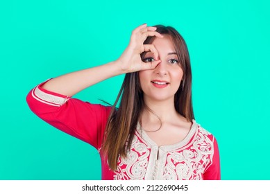 Young Beautiful Moroccan Woman Wearing Traditional Caftan Dress Over Green Background Doing Ok Gesture With Hand Smiling, Eye Looking Through Fingers With Happy Face.