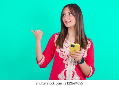 Young Beautiful Moroccan Woman Wearing Traditional Caftan Dress Over Green Background Using And Texting With Smartphone  Pointing And Showing With Thumb Up To The Side With Happy Face Smiling