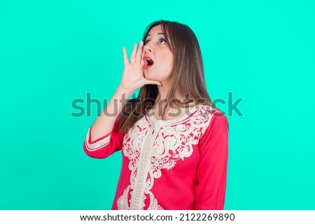 young beautiful moroccan woman wearing traditional caftan dress over green background shouting and screaming loud to side with hand on mouth. Communication concept.