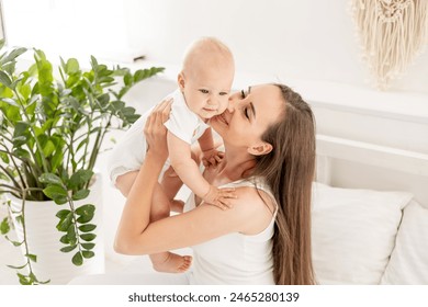 a young beautiful mother with a baby hugs and kisses him or plays with him, having fun on the bed in a bright bedroom, happy motherhood with maternity leave, maternal love and care. - Powered by Shutterstock