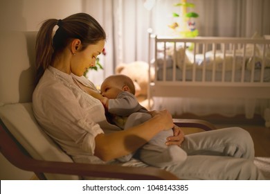 Young beautiful mother, breastfeeding her newborn baby boy at night, dim light. Mom breastfeeding infant