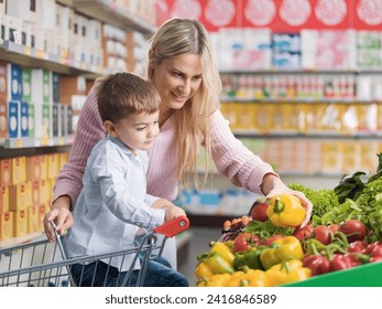 Young beautiful mother buying groceries with her son at the supermarket, they are picking healthy fresh vegetables in the produce section - Powered by Shutterstock