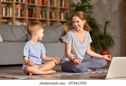 Young Beautiful Mother In Casual Wear Teaching Little Boy Son How To Meditate While Sitting Together In Lotus Pose On Floor In Living Room At Home, Happy Family Doing Yoga Exercises Online With Laptop