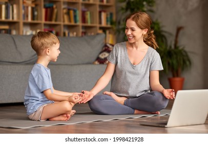 Young Beautiful Mother In Casual Wear Teaching Little Boy Son How To Meditate While Sitting Together In Lotus Pose On Floor In Living Room At Home, Happy Family Doing Yoga Exercises Online With Laptop