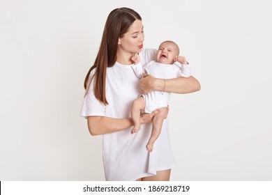 Young beautiful mother with cute little crying baby posing isolated over white background, mommy calm down her daughter, lady wearing casual t shirt, newborn in bodysuit.