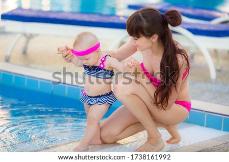 Young beautiful mother and daughter in bikini having fun resting near the pool. Happy family on summer vacation. Mom with baby girl sitting on the edge of the pool. Adventure, resort and rest concept.