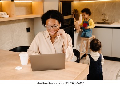 Young Beautiful Mother In Glasses And Headphones With Coffee Working With Laptop While Her Two Sons Playing Near In Cozy Kitchen