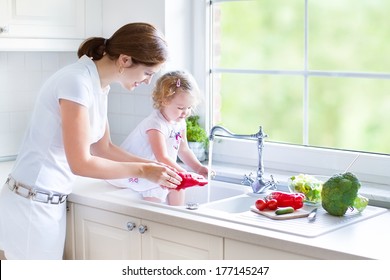 Young beautiful mother and her cute curly toddler daughter washing vegetables together in a kitchen sink getting ready to cook salad for lunch in a sunny white kitchen with a big garden view window - Powered by Shutterstock