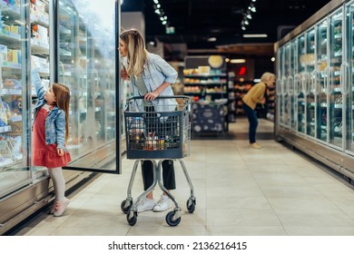 Young beautiful mother holding pushing shopping cart with her child in supermarket. Girl is choosing daily milk product picking up from shelf with her mother beside. Shopping for healthy. - Shutterstock ID 2136216415