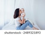 Young beautiful mother kisses her cute baby on cheek. White brunette touches her smiling child with her face. Pretty infant in a white bodysuit with his mom on a light background. Motherhood concept.