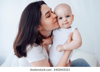 Young beautiful mother kisses her cute baby on cheek. White brunette touches her smiling child with her face. Pretty infant in a white bodysuit with his mom on a light background. Motherhood concept. - Powered by Shutterstock