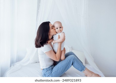 Young beautiful mother kisses her cute baby on cheek. White brunette touches her smiling child with her face. Pretty infant in a white bodysuit with his mom on a light background. Motherhood concept.