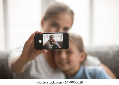 Young beautiful mother and little daughter sit on couch at home embracing making selfie view through mobile screen, younger and older sisters taking photo using modern wireless gadget concept image