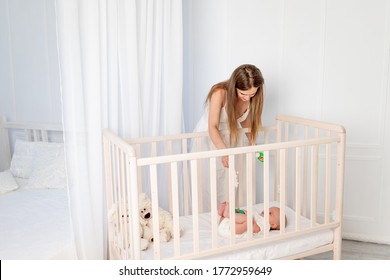 a young beautiful mother puts a 6-month-old baby in a crib, leaning over it in the nursery, mother's day, baby's morning, place for text