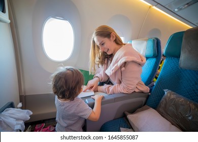 A Young Beautiful Mother Sits In An Airplane Chair And With Smile Shows With Finger A Page In A Book To Her Little Cute Toddler, Who Is Standing In Front Of Her. Top View. Copy Space, Blank Page