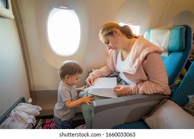 A Young Beautiful Mother Sits In An Airplane Chair And Reads A Book To Her Little Cute Toddler, Who Is Standing In Front Of Her. Close-up, Soft Focus, Top View.  Place For Copyspace, Empty Page