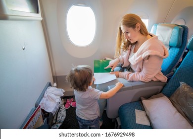 A Young Beautiful Mother Sits In An Airplane Chair And Shows With Finger A Page In A Book To Her Little Cute Toddler, Who Is Standing In Front Of Her. Top View. Copy Space, Blank Page