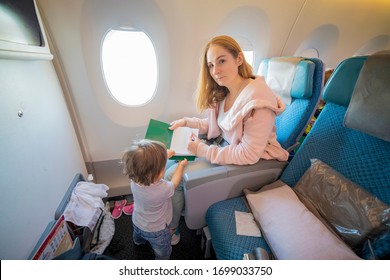 A Young Beautiful Mother Sits In An Airplane Chair Holds A Book And Shows It To The Camera, Her Little Cute Toddler Is Standing In Front Of Her. Top View. Copy Space, Blank Page