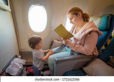 Young Beautiful Mother Sits In An Airplane Chair And Reads A Book. Her Little Cute Toddler Is Standing In Front Of Her. Close-up, Soft Focus, Top View. Place For Copyspace, Empty Blank Cover