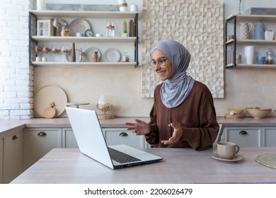 Young Beautiful Muslim Woman In Hijab Using Laptop For Video Call, Woman At Home Sitting In Kitchen Talking With Friends Remotely Smiling At Computer Web Camera.