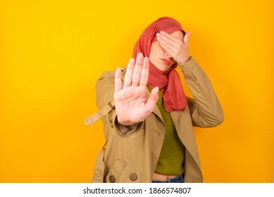 Young Beautiful Muslim Woman Wearing Hijab Against Yellow Background Covers Eyes With Palm And Doing Stop Gesture, Tries To Hide. Don't Look At Me, I Don't Want To See, Feels Ashamed Or Scared.