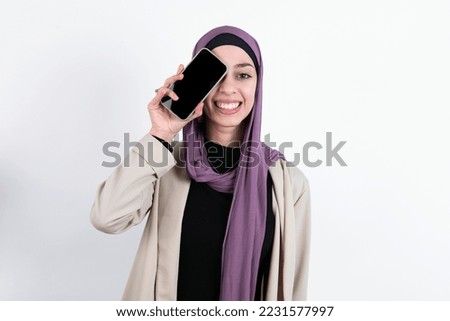 young beautiful muslim woman wearing hijab and jacket over white background  holding modern smartphone covering one eye while smiling