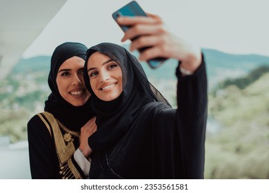 Young beautiful muslim women in fashionable dress with hijab using mobile phone while taking selfie picture on the balcony representing modern islam fashion technology and ramadan kareem concept