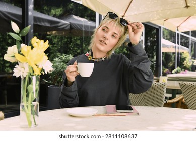 A young beautiful non binary person sits in a cafe, sipping coffee and embracing blissful vibes.