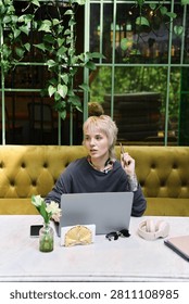 A young beautiful non binary person sits thoughtfully in a lively cafe, enjoying cozy ambiance.