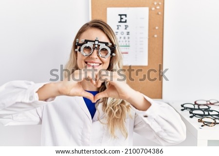 Young beautiful optician woman wearing optometry glasses at the clinic smiling in love showing heart symbol and shape with hands. romantic concept. 