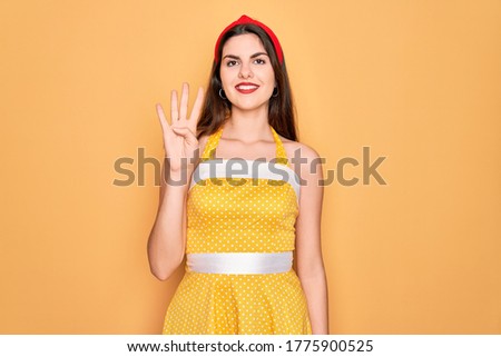 Young beautiful pin up woman wearing 50s fashion vintage dress over yellow background showing and pointing up with fingers number four while smiling confident and happy.