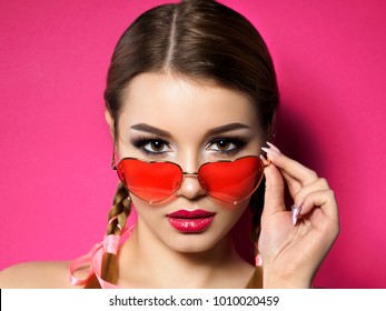 Young beautiful playful woman looks over her heart shaped red glasses. Valentines day, love or theme party concept. Smokey eyes and red lips makeup. Studio shot