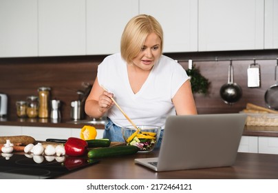 Young Beautiful Plus Size Plump Caucasian Woman Housewife Cooking Healthy Food, Making Vegetable Salad, Preparing Vegetarian Vegan Meal Using Laptop For Watching Culinary Video Recipe At Home Kitchen