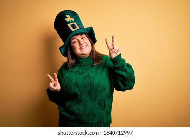 Young beautiful plus size woman wearing green hat with clover celebrating saint patricks day smiling looking to the camera showing fingers doing victory sign. Number two.