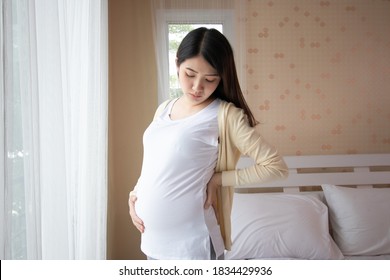 Young beautiful pregnant asian woman standing and touching belly and back near the window in bedroom. Pregnant women with low back pain, stress, anxiety concept. - Powered by Shutterstock