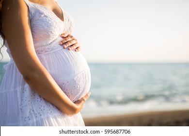 Young beautiful pregnant woman on the beach touching her belly with love and care making a heart shape. Relax by the calm sea in sunshine