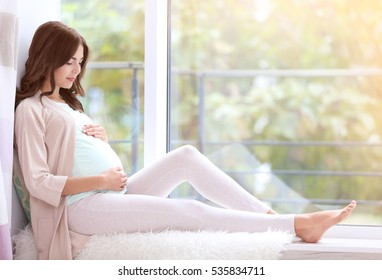 Young beautiful pregnant woman sitting on windowsill at home