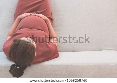 Young beautiful pregnant woman sitting on gray sofa and hand holding her belly in top view with copy space. Pregnancy, love, healthcare, motherhood, people, relaxation and expectation concept.