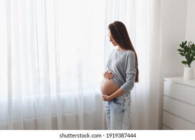 Young beautiful pregnant woman standing at home near the window.