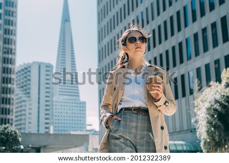 Young beautiful pretty girl walking along street in modern city skyscraper area with hand in pocket and holding cup of take away coffee. confident female office worker going to work on sunny morning