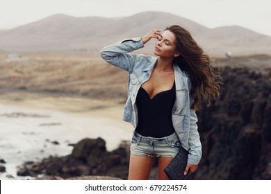 Young beautiful pretty stylish girl wearing jeans shorts and jacket walking on a windy day at rocky coastline. Feelings of freedom