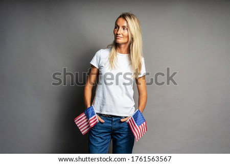 Young beautiful proud american blonde woman in white t-shirt and jeans with usa flags. Stylish portrait. Shining stars on the face. Independence. Patriot celebrates 4th of July. Copy space for text.