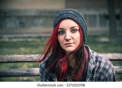 Young beautiful red hair girl sitting alone outdoors on the wooden bench with hat and shirt feeling anxious and depressed after she became a homeless person close up portrait 