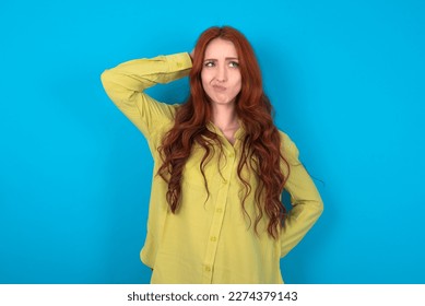 young beautiful red haired woman wearing green shirt over blue studio background confuse and wonder about question. Uncertain with doubt, thinking with hand on head. Pensive concept. - Powered by Shutterstock