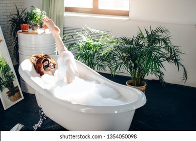 Young beautiful red-haired girl making a clay face mask for beauty during spa treatments in the bathroom, enjoying sunlight and warm water in bathtub, resort and vacation concept, summer time