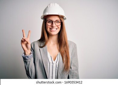 Young beautiful redhead architect woman wearing security helmet over white background smiling with happy face winking at the camera doing victory sign. Number two.