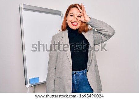 Young beautiful redhead businesswoman doing business presentation using magnetic board doing ok gesture with hand smiling, eye looking through fingers with happy face.