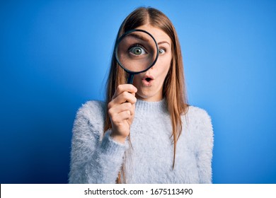 Young Beautiful Redhead Detective Woman Using Magnifying Glass Over Isolated Blue Background Scared In Shock With A Surprise Face, Afraid And Excited With Fear Expression