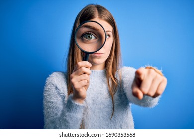 Young Beautiful Redhead Detective Woman Using Magnifying Glass Over Isolated Blue Background Pointing With Finger To The Camera And To You, Hand Sign, Positive And Confident Gesture From The Front